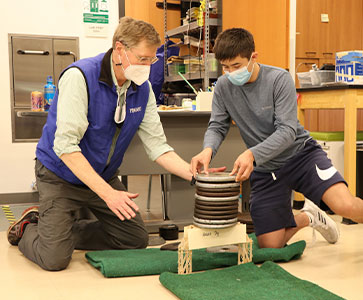 Teacher and student testing the construction of a wood bridge. Links to Closely Held Business Stock